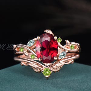 May include: Rose gold ring set featuring an oval-cut, deep red gemstone as the centerpiece. The ring is adorned with delicate leaf and vine details, accented by small green and blue gemstones. The design is intricate and nature-inspired.