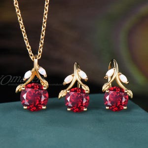 May include: A gold-toned jewelry set featuring a necklace and earrings. The necklace has a pendant with a large, round, red gemstone and two white accents. The earrings mirror the pendant design. The set is displayed on a teal surface.