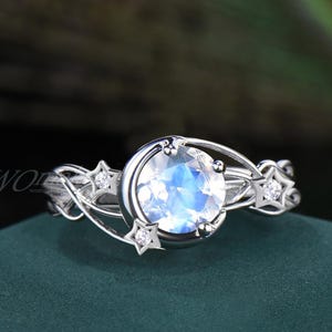 Könnte beinhalten: Ein silberner Ring mit einem großen, runden, irisierenden Edelstein, umrahmt von einem Mond-Design. Der Ring hat kleine, funkelnde Sternakzente und zarte Filigranarbeiten, die ein elegantes, himmlisches Thema schaffen.