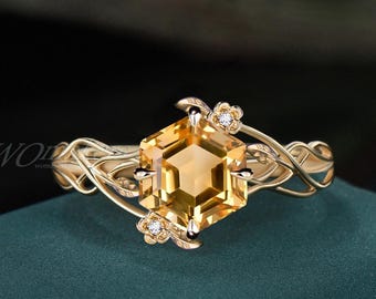 Könnte beinhalten: Ein goldener Ring mit einem großen, sechseckigen, gelben Edelstein als Mittelpunkt. Der Ring hat zarte, rankenartige Details und kleine Blumenakzente mit winzigen weißen Steinen. Der Ring ist vor einem dunkelgrünen Hintergrund platziert.