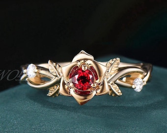 May include: A gold-colored ring featuring a rose design with a central red gemstone. The ring has delicate leaf accents and two smaller, clear gemstones on either side of the rose. The ring is set against a dark green background.