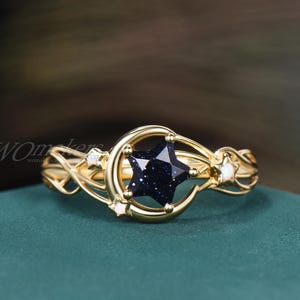 May include: A gold-colored ring featuring a dark blue star-shaped stone set within a crescent moon design. The ring has delicate, swirling details and small, round accent stones. The ring is displayed on a green surface.