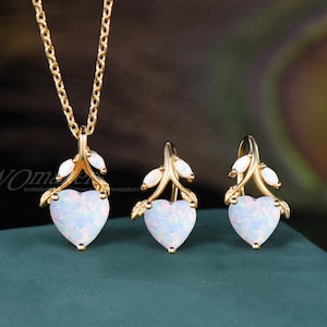 May include: A gold-coloured jewellery set comprising a necklace and earrings. Each piece features a heart-shaped, opalescent pendant. The pendants are accented with gold-coloured leaves and small white stones. The necklace chain is also gold-coloured.