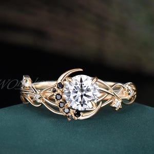 May include: A gold ring featuring a large, round, white gemstone at the center. The ring is adorned with crescent moon and star designs, accented with small black gemstones. The band has intricate, flowing details.