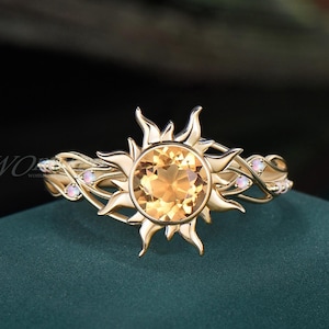 May include: A gold ring featuring a sunburst design with a large, round, yellow gemstone at the center. The band has a delicate, swirling pattern with small, white gemstones. The ring is set against a dark green background.