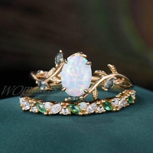 May include: A gold ring set with a large oval opal and smaller green gemstones. The ring features a delicate branch design with leaves. The ring is paired with a matching band featuring smaller opals and green gemstones.