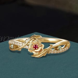 May include: A gold-colored ring featuring a floral design with a red gemstone center and small clear and green gemstones. The ring has a delicate, nature-inspired aesthetic with a floral and vine motif.
