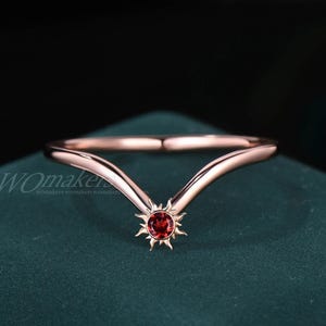 Könnte beinhalten: Roségoldener Ring mit Sonnenmuster und einem runden, roten Edelstein. Der Ring hat ein gebogenes Band und eine sonnenförmige Fassung. Der Ring wird auf einer dunkelgrünen Oberfläche präsentiert.