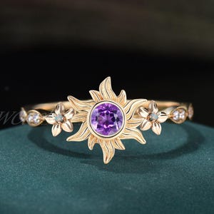 May include: A gold ring featuring a central purple gemstone surrounded by a sunburst design. The ring is adorned with small flower and gemstone accents on the band. The ring is set against a dark green background.