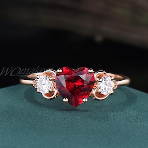May include: Rose gold ring featuring a heart-shaped, deep red gemstone as the centerpiece, flanked by two smaller, round, white gemstones. The ring's band and setting have a delicate, ornate design, set against a dark green background.