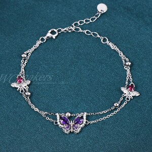 May include: A silver bracelet featuring three butterfly charms. The central butterfly is purple, while the other two are pink. The bracelet has a delicate chain design and is set against a teal background.