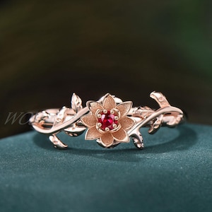May include: Rose gold ring featuring a floral design with a central red gemstone. The ring's band is adorned with delicate leaves and vines, creating an elegant and nature-inspired aesthetic. The ring is a piece of jewelry.