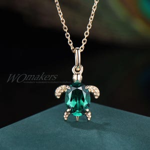 May include: A gold-colored necklace featuring a turtle pendant. The pendant has a large, oval-shaped, emerald-green gemstone body, with gold-colored accents on the flippers and head. The necklace chain is also gold-colored.