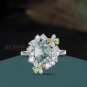May include: A silver ring featuring an oval-cut, moss agate gemstone. The ring is adorned with clusters of small, clear gemstones and accented with yellow and green gemstones in a floral design. The ring is set against a dark teal background.