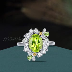 May include: A silver ring featuring a large, oval-cut peridot gemstone as the centerpiece. The peridot is surrounded by smaller white and yellow gemstones, and green flower-shaped accents. The ring has a delicate, nature-inspired design.