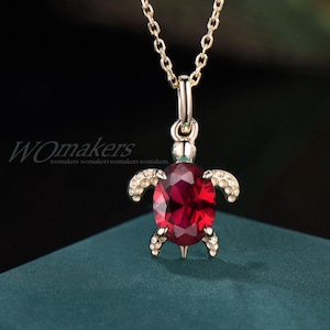 May include: A gold-toned necklace featuring a turtle-shaped pendant with a large, faceted, oval-cut deep red gemstone as the shell. The turtle's head and four flippers are accented with small, clear, sparkling stones. The text WOmakers is visible.