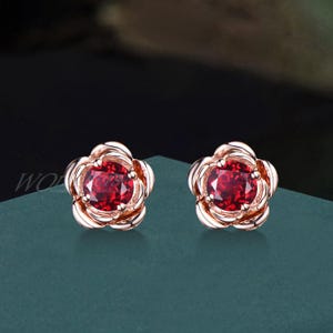 May include: A pair of rose gold stud earrings with a red gemstone in the center of each flower-shaped setting.