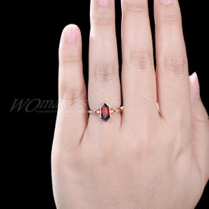 K&ouml;nnte beinhalten: Ein Ring aus Ros&eacute;gold mit einem roten Edelstein, der in eine geometrische Form gefasst ist. Der Ring befindet sich an einem Finger.