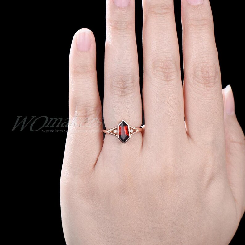 K&ouml;nnte beinhalten: Ein Ring aus Ros&eacute;gold mit einem roten Edelstein, der in eine geometrische Form gefasst ist. Der Ring befindet sich an einem Finger.