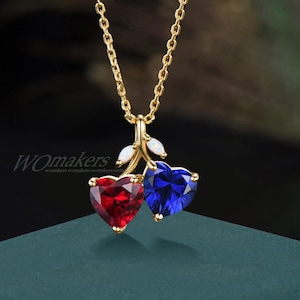Puede incluir: Un collar dorado con un colgante de cereza. El colgante presenta dos piedras preciosas en forma de corazón, una roja y otra azul, con pequeños detalles blancos. El collar se muestra sobre una superficie verde azulado oscuro.