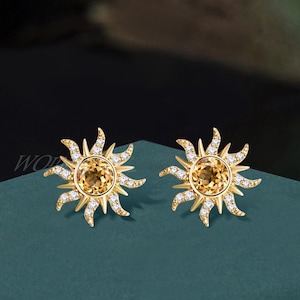 May include: A pair of gold stud earrings shaped like suns, each featuring a round, faceted yellow citrine gemstone at the center surrounded by radiating rays encrusted with small, clear, sparkling white stones in a pavé setting.