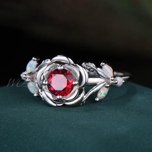 May include: A silver ring with a red gemstone set in a rose-shaped design. The ring is adorned with white opal accents on the sides.