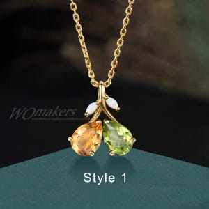 Könnte beinhalten: Eine goldene Kettenkette mit einem Anhänger, der zwei birnenförmige Edelsteine ​​​​zeigt, einen grünen und einen orangefarbenen, mit kleinen weißen Opal-Akzenten. Der Anhänger hat die Form eines Zweigs mit Blättern. Der Text "Style 1" ist unterhalb des Anhängers sichtbar.