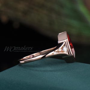 K&ouml;nnte beinhalten: Ein Ring aus Ros&eacute;gold mit einem gro&szlig;en roten Edelstein und drei kleineren wei&szlig;en Edelsteinen. Der Ring liegt auf einer gr&uuml;nen Oberfl&auml;che.