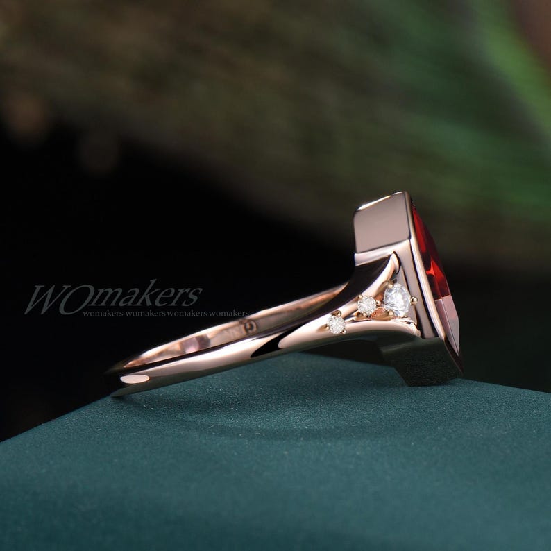 K&ouml;nnte beinhalten: Ein Ring aus Ros&eacute;gold mit einem gro&szlig;en roten Edelstein und drei kleineren wei&szlig;en Edelsteinen. Der Ring liegt auf einer gr&uuml;nen Oberfl&auml;che.