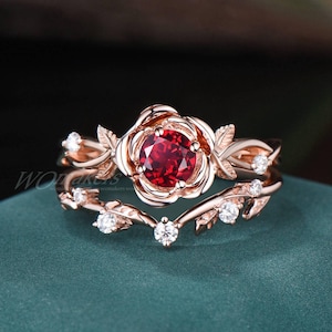 May include: A rose gold ring set with a large red gemstone surrounded by smaller clear gemstones. The ring features a delicate rose design with leaves and vines.