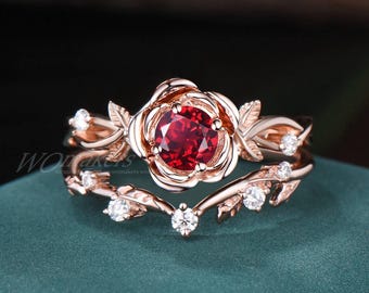Könnte beinhalten: Ein Ring aus Roségold mit einem großen roten Edelstein, der von kleineren klaren Edelsteinen umgeben ist. Der Ring hat ein zartes Rosenmotiv mit Blättern und Ranken.