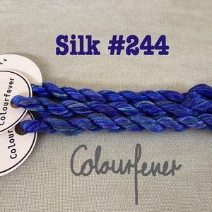 Op de afbeelding: Drie klossen blauwe zijden borduurgaren met een wit label waarop "Colourfever" en het nummer "244" staat. Het label is met een klein gaatje aan het garen bevestigd.