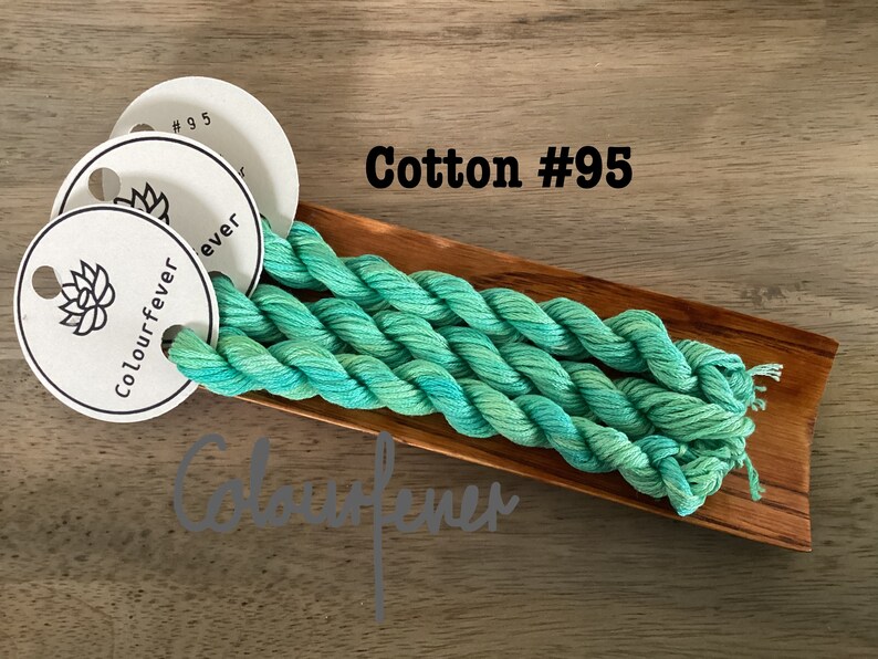 Puede incluir: Tres madejas de hilo de bordar de algod&oacute;n verde azulado y verde en una bandeja de madera. El hilo est&aacute; etiquetado como "Colourfever #95".