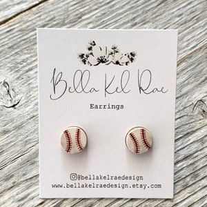 May include: A pair of baseball stud earrings on a white card with the text "Bella Kel Rae Earrings". The earrings are round with a baseball design, featuring red stitching on a cream background. The card also includes social media and website information.