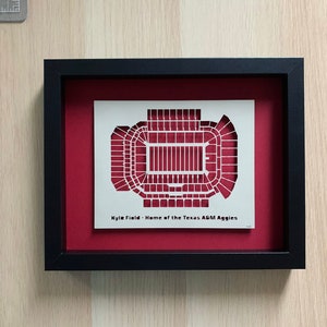 May include: A framed black and white paper cut-out of a football stadium with the text "Kyle Field - Home of the Texas A&M Aggies". The frame is black with a maroon mat.