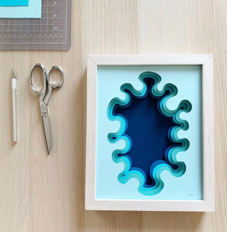 May include: A framed paper art piece with a blue and teal layered design. The design is a series of concentric circles with a wavy edge, creating a 3D effect. The frame is light wood.