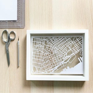 May include: A white paper cutout of a city map in a white frame. The map is detailed and shows streets and waterways.