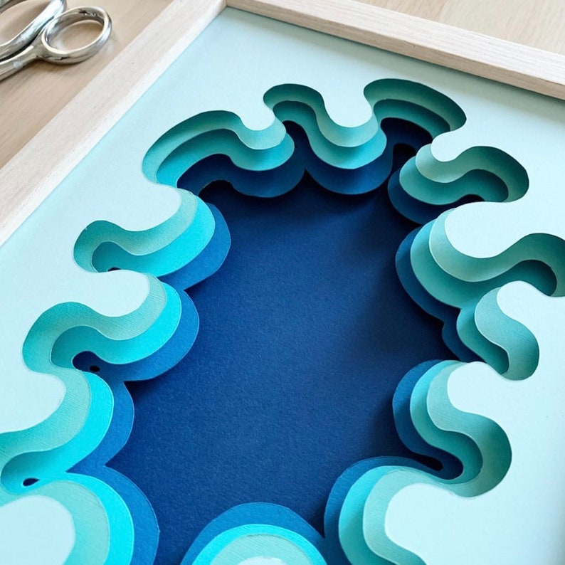 May include: A framed paper art piece with a blue and white layered design that resembles waves. The waves are cut out of the paper and layered to create a 3D effect.