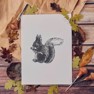 May include: A black and white illustration of a squirrel holding a nut, printed on a white card. The card is surrounded by brown paper and fall leaves.
