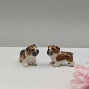 May include: Two small ceramic bulldog figurines, one brown and white, the other brown and white, facing each other on a white surface.