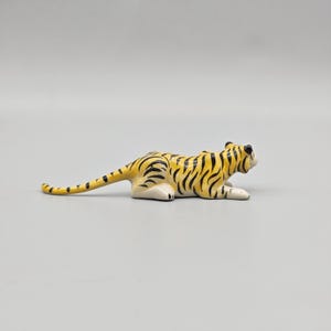 May include: A small, ceramic tiger figurine in a resting pose. The tiger has a yellow body with black stripes, white paws, and a long tail. The figurine is on a plain gray background.