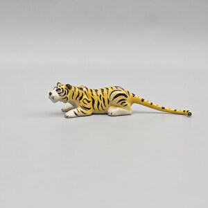 May include: A small, yellow and black toy tiger in a crouching position. The tiger has black stripes and a white face and paws. The tail is long and thin, with black spots. The toy is made of a hard material.