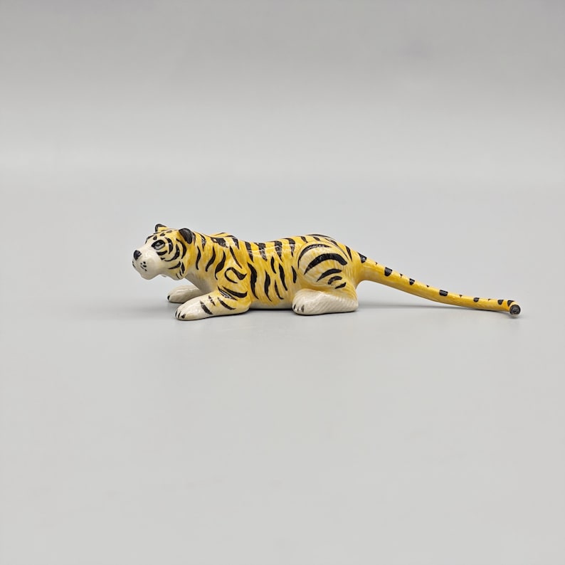 May include: A small, yellow and black toy tiger in a crouching position. The tiger has black stripes and a white face and paws. The tail is long and thin, with black spots. The toy is made of a hard material.