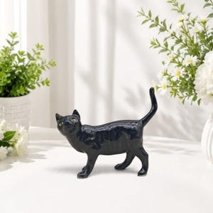 May include: A glossy black ceramic cat figurine standing in a prowling pose with its tail held high. The cat features pointed ears and yellow eyes, designed as a functional holder for rings or other small jewelry items on its tail.