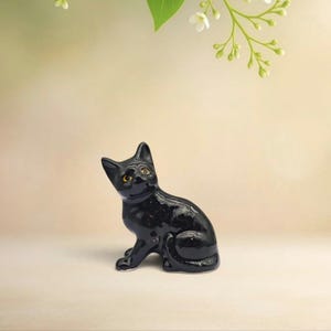 Puede incluir: Una pequeña figura de gato de cerámica negra brillante en posición sentada. El gato tiene orejas puntiagudas, una superficie lisa y reflectante, y ojos de color amarillo anaranjado que miran hacia arriba. La figura tiene un acabado oscuro.