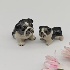May include: Two ceramic bulldog figurines, one larger than the other, both with black and white markings. The larger bulldog is sitting, while the smaller one is standing. Both figurines have a realistic appearance.