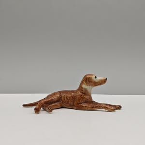 May include: A ceramic figurine of a dog in a resting pose. The dog is primarily brown with lighter brown and white accents on the face and chest. The figurine is on a white surface against a gray background.