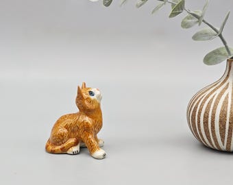May include: A ceramic figurine of a ginger cat with blue eyes, looking upwards. The cat is sitting with its front paws extended. A decorative vase with vertical brown stripes and a sprig of greenery are in the background.