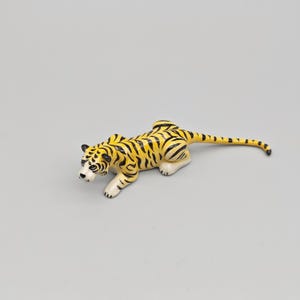 May include: A ceramic tiger figurine in a crouching pose. The tiger has a yellow body with black stripes, white paws, and a white face with black markings. The tail is long and striped, and the ears are black.
