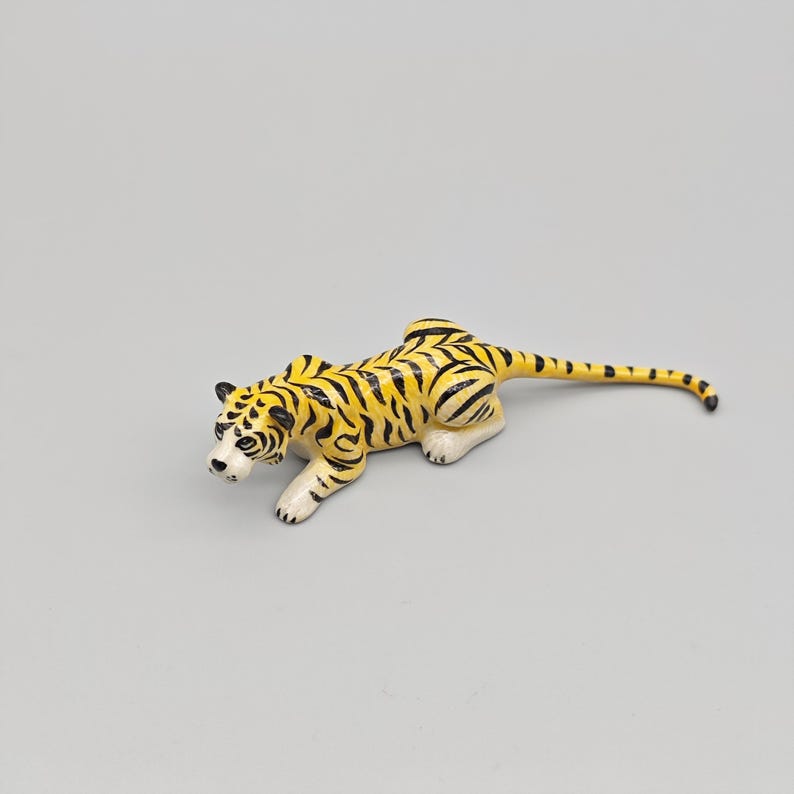 May include: A ceramic tiger figurine in a crouching pose. The tiger has a yellow body with black stripes, white paws, and a white face with black markings. The tail is long and striped, and the ears are black.